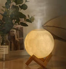 Real photo: Luminous, warm white moon-shaped aroma diffuser lamp with textured craters, on a light brown wooden stand. Mist gently rises from the top, in a cozy home setting.