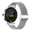 Back of NX23 Pro Smartwatch: silver metallic casing, Milanese mesh band. Black sensor panel with glowing green heart rate & pedometer sensors. Text: WATER RESISTANT, INPUT:5V-1A, CE, FC. Real product photo.