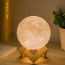 Cream-white, textured moon lamp and aroma diffuser glowing warmly on a light brown wooden stand. It's on a desk with books and a small plant, providing a cozy atmosphere.