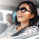 Smiling woman in a car wearing stylish black sunglasses with slightly thick temples and dark, reflective lenses. The design hints at integrated audio. Real photo.
