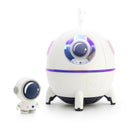 White plastic spaceship humidifier with glowing purple/blue LED window, astronaut figure inside, and mist rising. Stands on black legs, beside a small white astronaut figurine. Studio product shot.