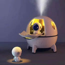 Spaceship Explorer Humidifier: a white, spherical device with tripod legs. An astronaut sits inside an open, yellow-lit hatch as mist streams from the top. A second astronaut watches from below. Playful 3-in-1 product image.
