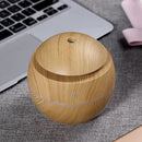Round, light wood grain aroma diffuser with a translucent band and power symbol, on a grey mat next to a laptop. Modern, compact design.