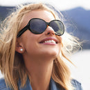 A blonde woman with windblown hair smiles, wearing stylish black, rounded Gafas Sol Bluetooth F07 sunglasses with dark lenses. She's outdoors in a real photo.