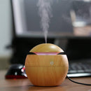 Wood grain spherical aroma diffuser emitting mist, with a pink LED glow, on a wooden desk. Features a power icon. Real photo of a multifunctional humidifier for a fresh environment.