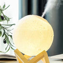 Glowing moon aroma diffuser lamp, off-white with crater texture, emitting mist. It stands on a light brown wooden base. Realistic home decor piece.