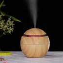 Spherical wood-grain aroma diffuser, light brown with reddish lines, emitting mist. Features a glowing pink light ring and power button, on a white textured table. Real image.