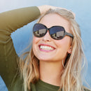 Smiling blonde woman wearing stylish black Bluetooth F07 sunglasses with thick frames and dark, reflective lenses. She has a green top and is against a bright blue wall.