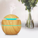 Multifunctional wood-grain aroma diffuser, light brown with teal LED glow & power button, emitting white mist. Spherical design on a white surface. Real product photo for fresh ambiance.