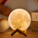 Glowing 3D moon lamp with realistic white and yellow crater texture, resting on a minimalist dark brown wooden stand, emitting a warm light. Real product photo.