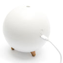 Minimalist white spherical humidifier/aroma diffuser with three light wooden legs, shown charging on a white background. Features a top mist vent and a power button icon next to the charging port. Real product photo.