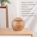Round light wood grain aroma diffuser, mist rising, on a wooden table. Features listed in Spanish: "Difusor aromático, Apagado automático, Silencioso, Filtro removible, Antibacterial, Color madera clara, Capacidad de 130 ml".