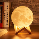 A glowing yellow 3D moon lamp with crater textures sits on a minimalist dark wood stand. Real photo on a wooden surface with books, one labeled "BROOKLYN'S FINEST".