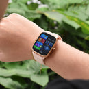 SmartWatch KT65 Pro with rose gold bezel & blush pink silicone band on a wrist, alongside a gold chain bracelet. The vibrant digital display shows time 12:02, date, 20°C, steps, and fitness metrics. Real photo, green foliage background.