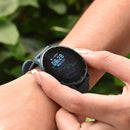 SmartWatch ZL02 Pro on a wrist. Round black screen shows 08:28, WED 03/01, and heart rate icon. Features a dark grey silicone strap and silver bezel. A hand touches the side. Real photo with a green background.