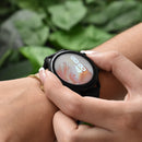 Woman's hand adjusts a black Smartwatch ZL73J on her wrist. Its round digital screen shows '8:48 JAN 03' with a soft pink/white abstract watch face. A gold bracelet is on her wrist, with green foliage background. Real photo.