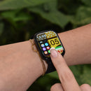 Black rectangular smartwatch on a wrist, displaying 12:05, app icons, and a burger-themed background. A finger touches its screen. A gold chain bracelet is on the wrist. Real image with blurred green foliage.