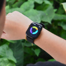 Black square ZL99J Reloj Inteligente on a wrist, featuring gold lugs & a black strap. The screen displays colorful activity rings and 12:07. Real image against blurred green foliage, showcasing its water-resistant build.