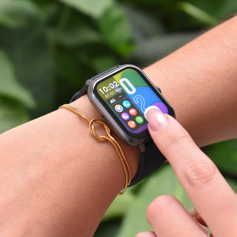 Dark grey ZL54CJ smartwatch with black band on a wrist. A finger touches the vibrant screen displaying 10:02, 01-01 Mon, and app icons. A gold knot bracelet is also visible. Real photo outdoors.