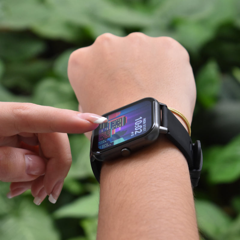 Hand interacts with ZL54CJ smartwatch: black silicone band, dark gray rectangular frame. Color screen shows 10:02, MON 01/01, city graphic. Real photo against green foliage, highlighting the modern 'Reloj Inteligente'.