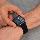 Close-up of a person's wrist wearing a dark grey SmartWatch KT65 Pro with a black silicone strap. A finger touches its vibrant screen showing time (14:02), weather (20°C), steps, and app icons. Real photo.