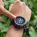 Hand touching a black, rugged Smartwatch LC16 with a silver bezel. Its round screen shows colorful app icons. "SPORT WATCH" is etched on the bezel, set against green foliage.