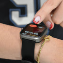 Real photo of a Smartwatch NX22 with a black strap & gray body on a wrist. A finger with red nails taps the screen showing "Cronómetro", "00:10.50", "04:57", and red buttons "Detener" & "Vuelta". A gold bracelet is also on the wrist.