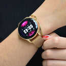 Rose gold NX23 Pro smartwatch on a woman's wrist, with a metallic mesh band. The round digital display shows 00:43, Sun 01-16, 20°C, and vibrant pink lotus flower graphics. Real product photo.