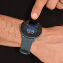 SmartWatch ZL02 Pro on a hairy wrist, touched by a finger. The round black screen displays 10:05, SAT 2701, --BPM, and a dynamic blue/green wave graphic. Features a teal silicone band. Real photo.