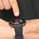 Real photo: Smartwatch NX18 with black metallic bezel and silicone strap on a wrist. A finger taps its round screen displaying 'Settings', 'Watch Faces', 'Brightness'. Bezel shows compass directions.