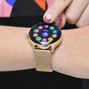 NX23 Pro Smartwatch in rose gold/gold, with a round black touchscreen displaying colorful app icons including 'Sueño' (Sleep). Worn on a woman's wrist, featuring a metallic mesh strap and a finger interacting with the screen. Real image.