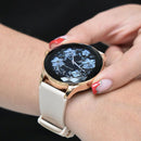 NX23 Pro smartwatch mujer: rose gold faceted case, cream silicone strap. Black screen shows blue floral design, 'Smart Watch', '01', and crown 'V' logo. Worn on a woman's wrist. Real photo.