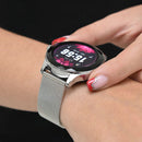 NX23 Pro Smartwatch Mujer on a wrist, with a silver metal mesh strap and textured round bezel. The black screen shows 15:56, Mon 01-03, and 20°C on a pink background. Real, stylish photo.