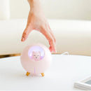 Real photo of a pink spherical "Planet Cat" humidifier on wooden legs, featuring a clear window with a glowing, smiling cat night light inside. A hand reaches toward it on a light table.