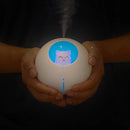 White spherical Planet Cat humidifier held in hands, emitting mist. It features a blue glowing cat face night light and a small blue indicator light below it, against a dark backdrop. Real photo.