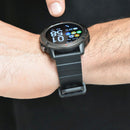 SmartWatch NX9, a real photo on a wrist. Features dark gray rugged bezel, black strap, and round Amoled screen showing 1:58, Sat 01, 20°C with app icons. A finger touches the display.