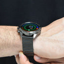NX1 Pro smartwatch on a male wrist. Features a dark gunmetal gray metal case & link bracelet. The round digital screen shows colorful concentric data rings and white numbers. A red accent button is visible. Real photo, conveying a sleek, robust design.