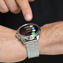 Elegant silver NX1 Pro smartwatch with a metallic linked band on a wrist. A finger taps the black screen showing colorful call, contacts, and call log options in Spanish. Real photo.
