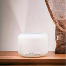 White V-M500 ultrasonic humidifier with mist emerging, shown on a wooden surface. Features tactile buttons labeled 'LIGHT', 'HIGH/LOW', 'MIST' and timer settings (60, 120, 180, ON). A real image with a modern, clean design and a brown textured fabric in the background.